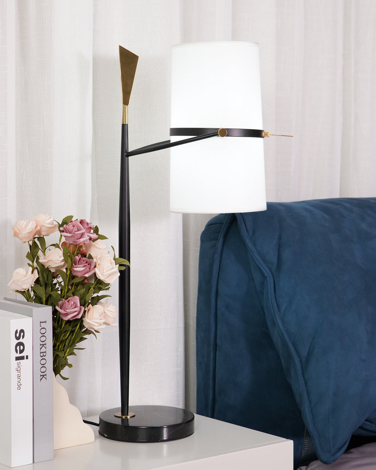Straight-on view of this Clérachic black table lamp, the white shade held level and the gold tip catching light, beside roses and stacked books.