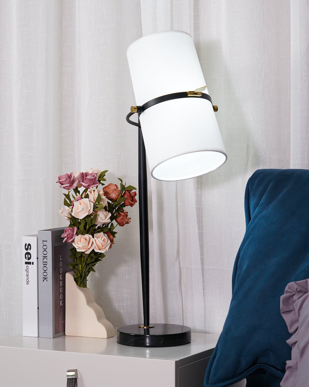 Side view of this Clérachic lamp's tilted white shade and black stem on a round black base, with pink roses and books at the left.