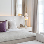 Bedroom with a Clérachic table lamp glowing softly on a white nightstand beside a bed with purple and grey cushions and tall curtains.