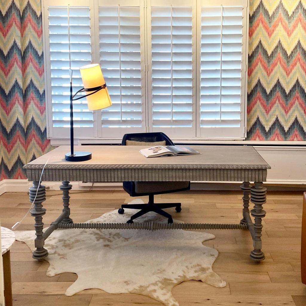 A Clérachic table lamp lit on a grey writing desk in front of white shutters and a zigzag-patterned wall, with a cowhide rug below.