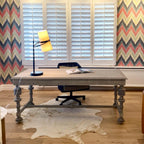 A Clérachic table lamp lit on a grey writing desk in front of white shutters and a zigzag-patterned wall, with a cowhide rug below.
