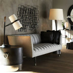 A Clérachic table lamp on a round black side table beside a grey channel-tufted sofa, with a metal wall sculpture behind it.