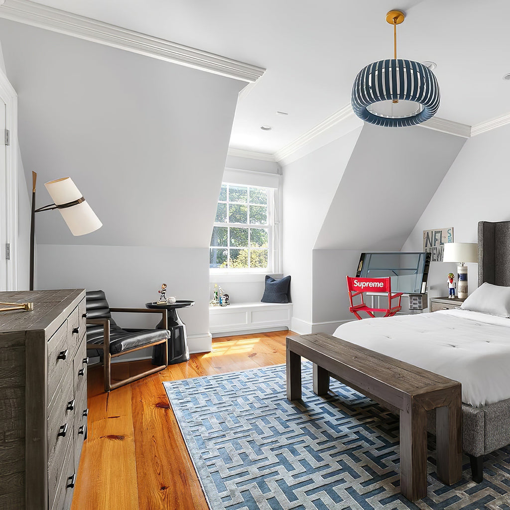 Attic bedroom with a Clérachic table lamp on a dresser beside a window seat, a blue patterned rug and a bench at the foot of the bed.