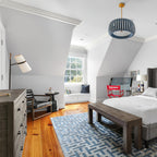Attic bedroom with a Clérachic table lamp on a dresser beside a window seat, a blue patterned rug and a bench at the foot of the bed.