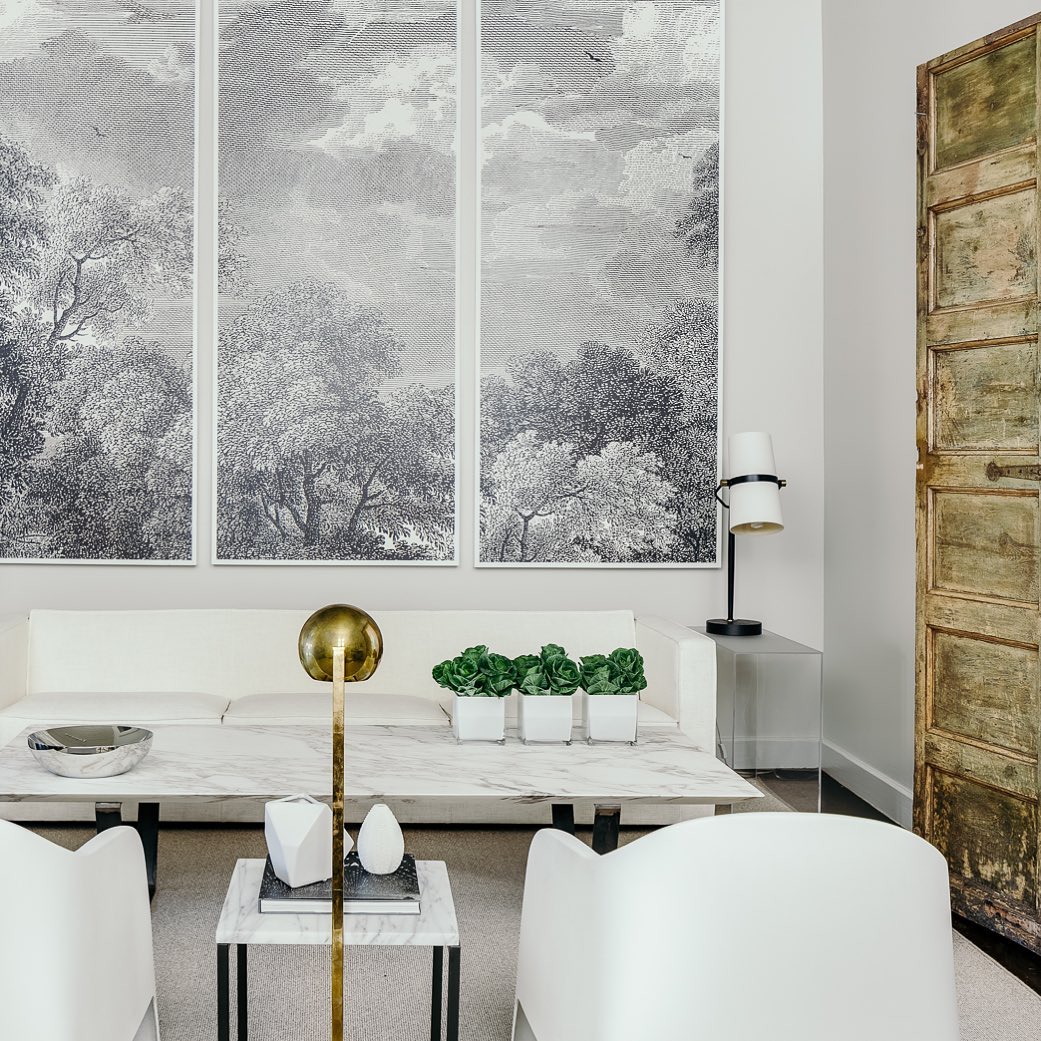 Pale living room with a small Clérachic table lamp on a glass side table at right, below tall grey landscape panels and a weathered door.