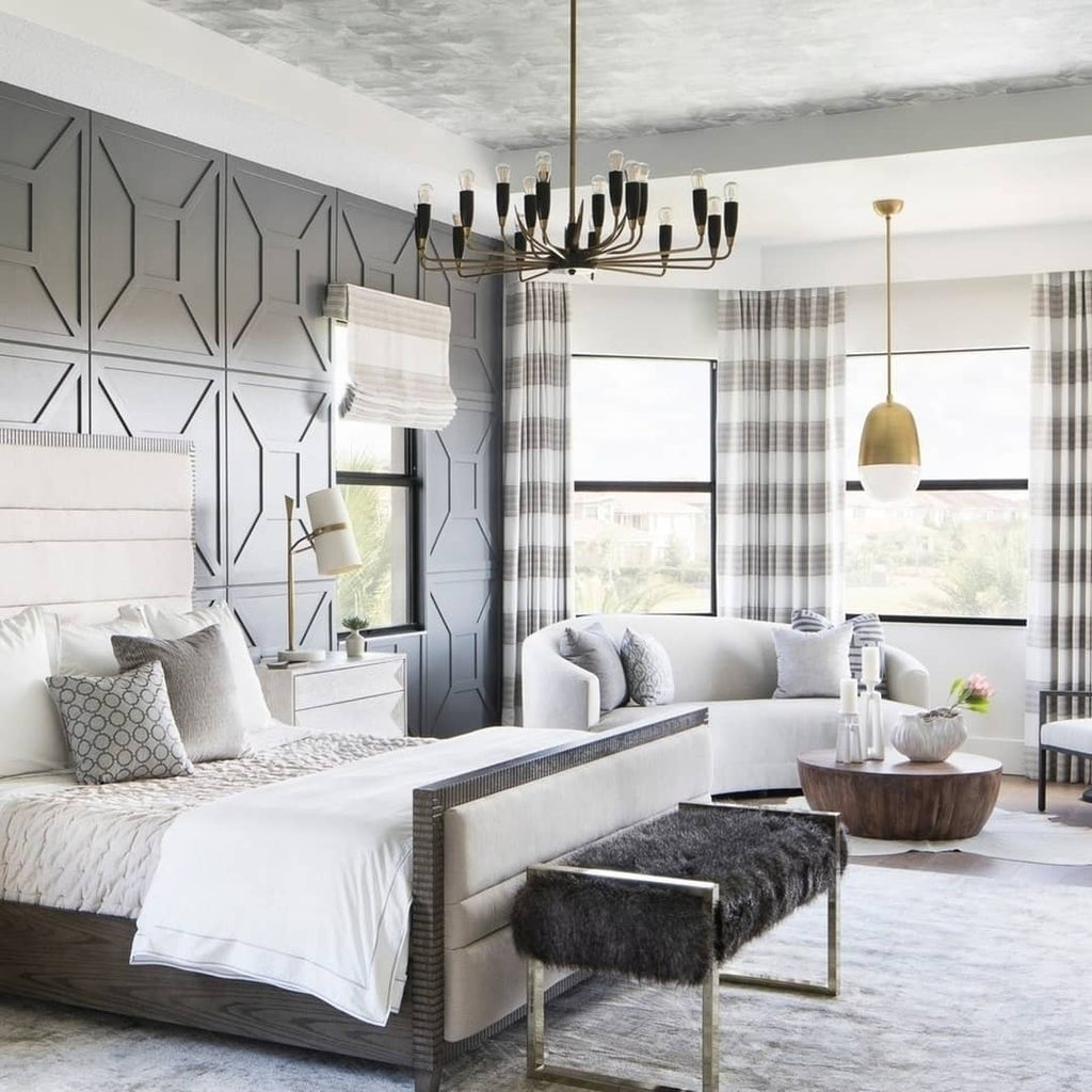 Large bedroom with a Clérachic table lamp on a bedside table, a tufted grey bed, a black multi-arm chandelier and a bay window.