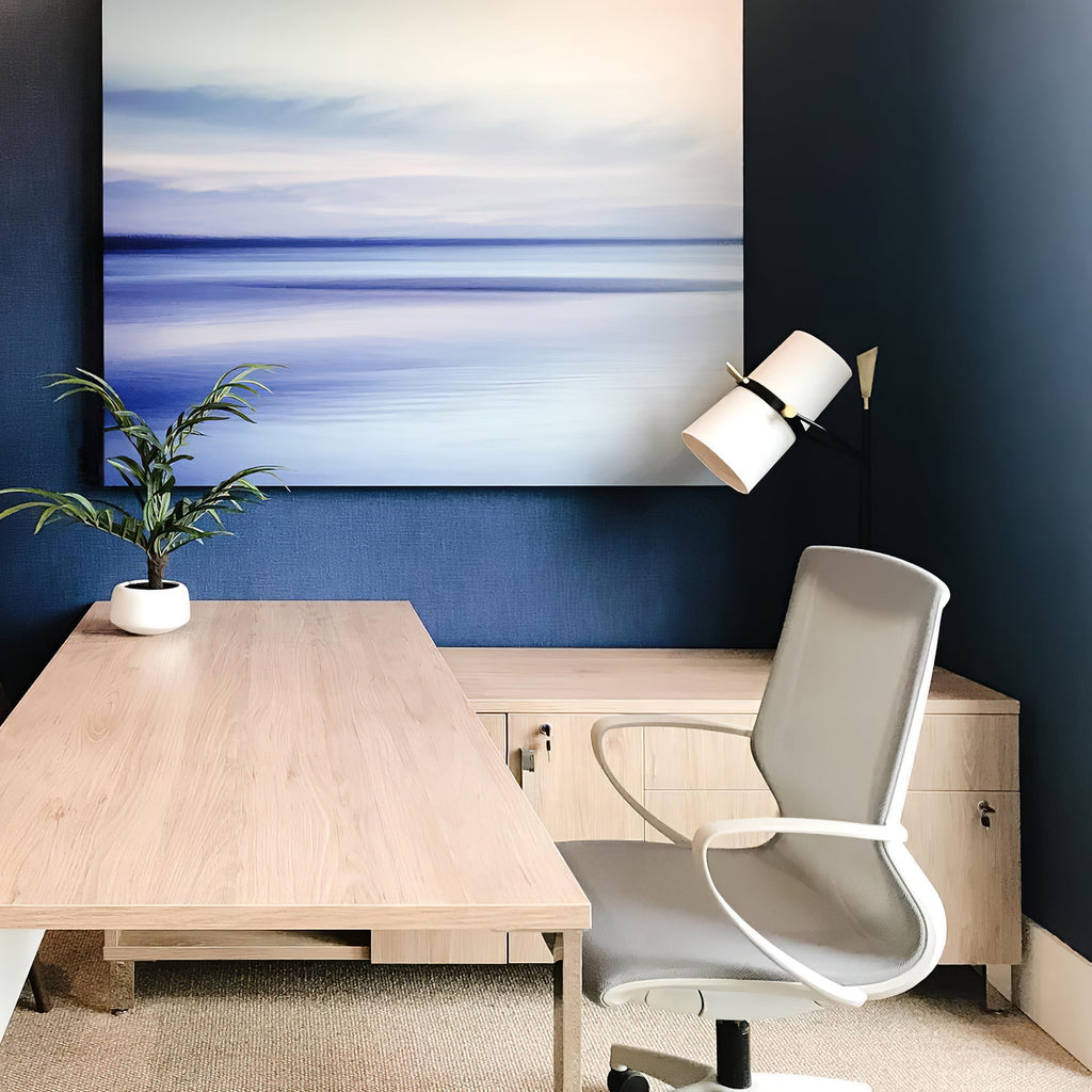 A Clérachic table lamp at the edge of a navy-painted wall beside a blue seascape canvas, over a light wood desk and grey chair.