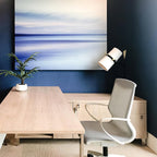 A Clérachic table lamp at the edge of a navy-painted wall beside a blue seascape canvas, over a light wood desk and grey chair.