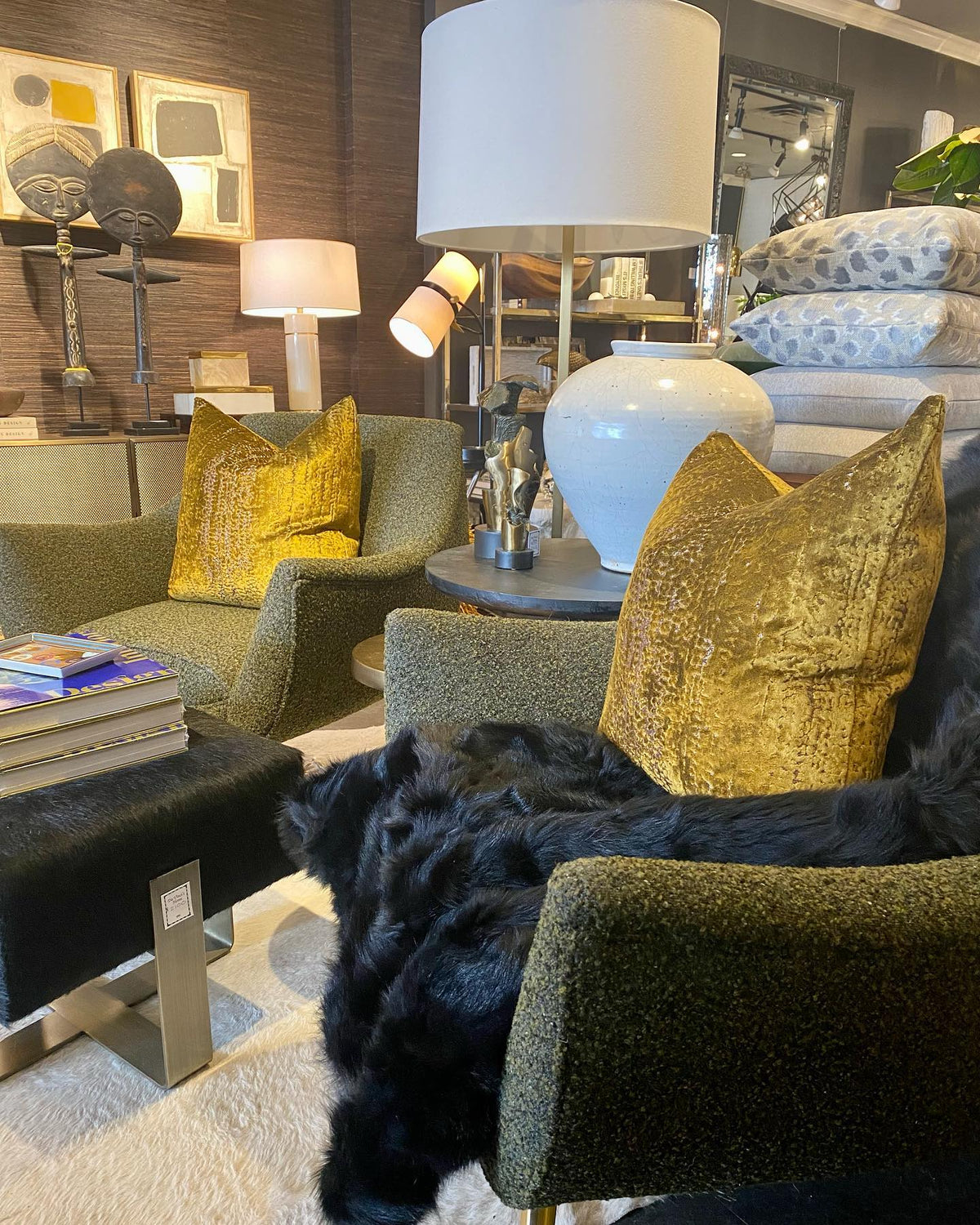 Showroom vignette with a Clérachic table lamp behind olive velvet chairs and gold cushions, near a white ceramic lamp and a black fur throw.