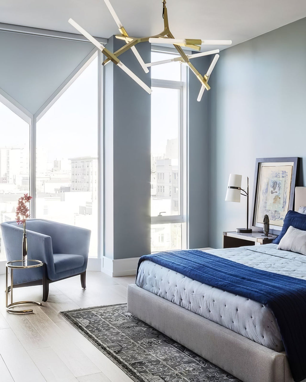 Blue-grey bedroom with a Clérachic table lamp on the bedside table, a bed with a navy throw, an armchair and a gold branching chandelier above.