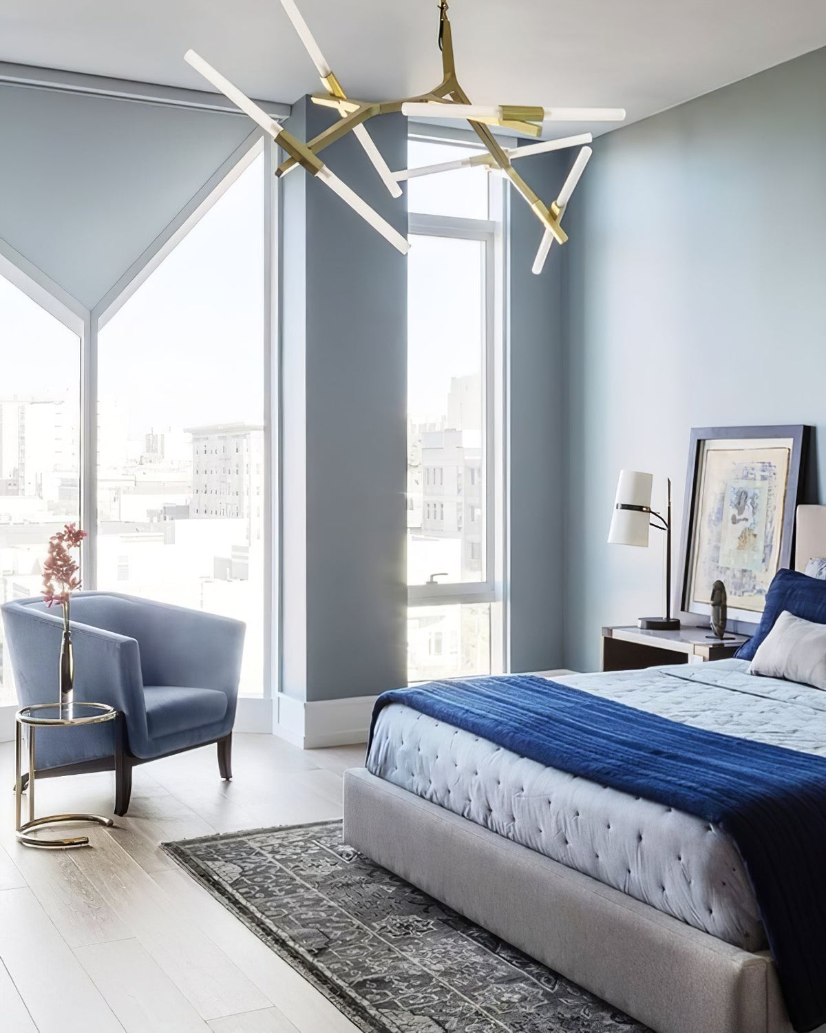 Blue-grey bedroom with a Clérachic table lamp on the bedside table, a bed with a navy throw, an armchair and a gold branching chandelier above.