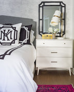 Close view of a Clérachic table lamp on a white chest of drawers beside a black-framed mirror, with monogrammed black and white bedding.