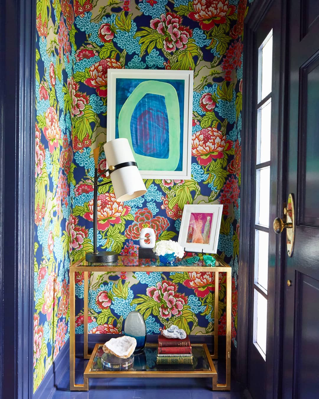 Hallway nook with a Clérachic table lamp on a gold glass side table against bold floral wallpaper and a green framed print.