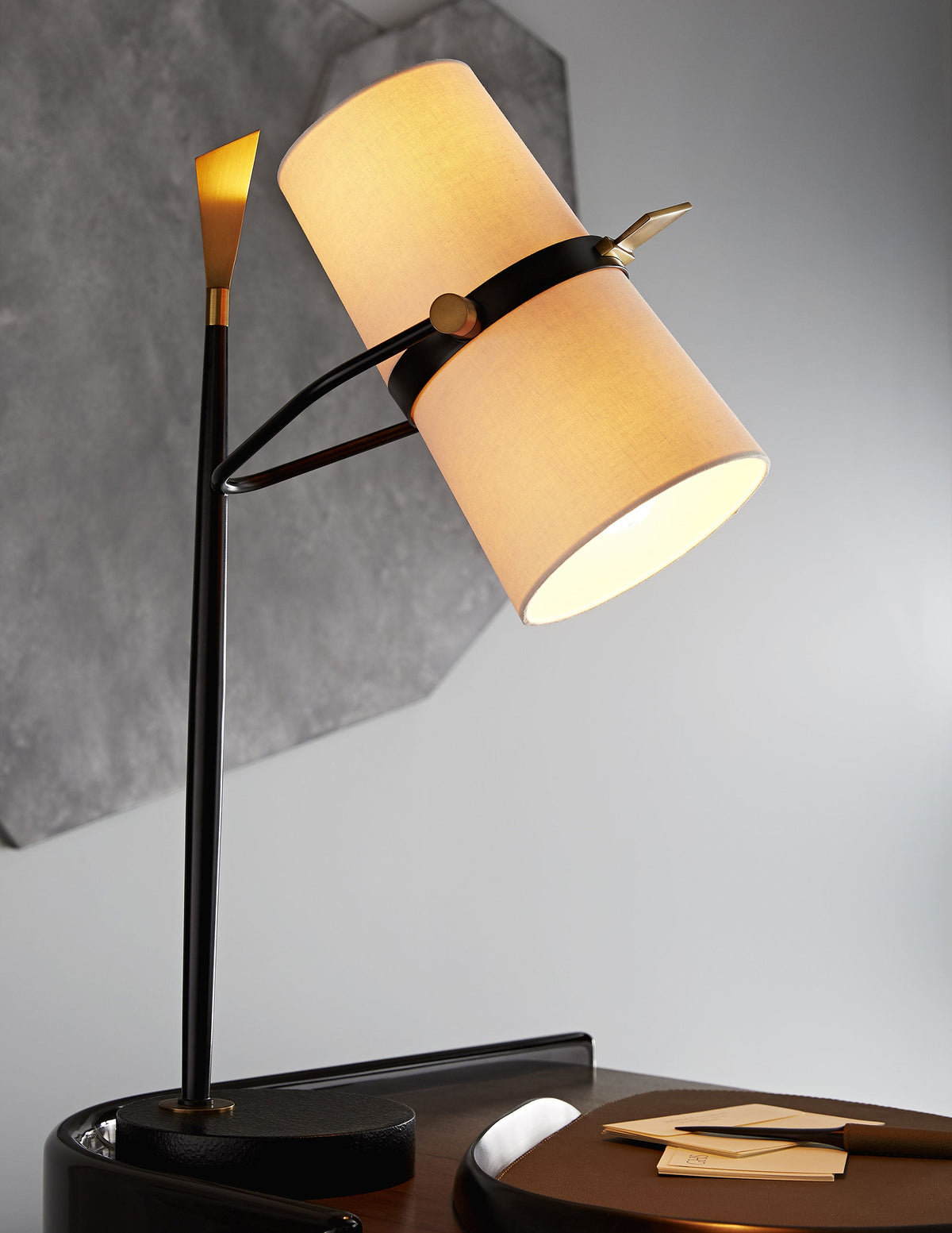 Close view of this Clérachic table lamp, its black stem and tilted shade glowing warm amber on a dark base against a pale grey wall.
