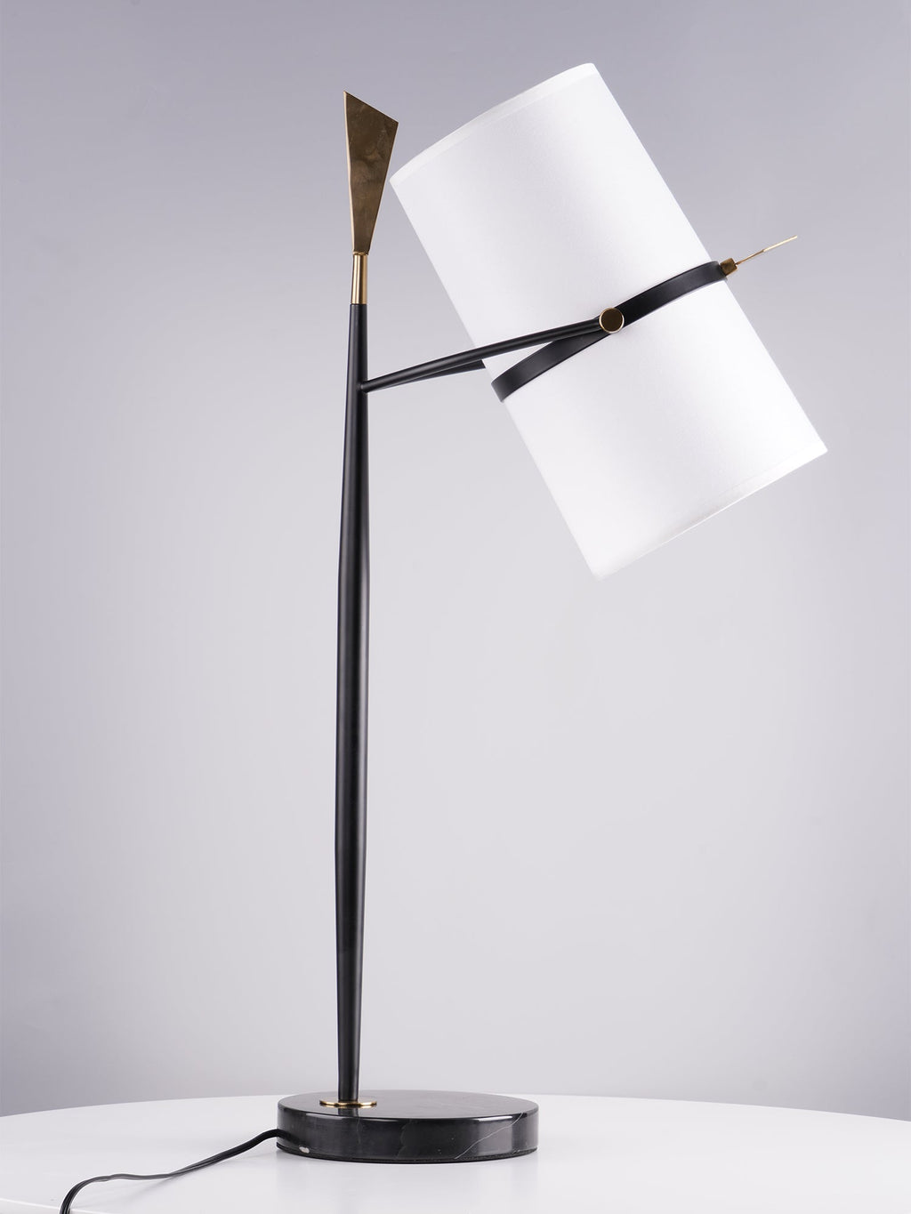 Black-stemmed Clérachic table lamp with a black marble base on a white round table, the white shade tilted right against a grey backdrop.