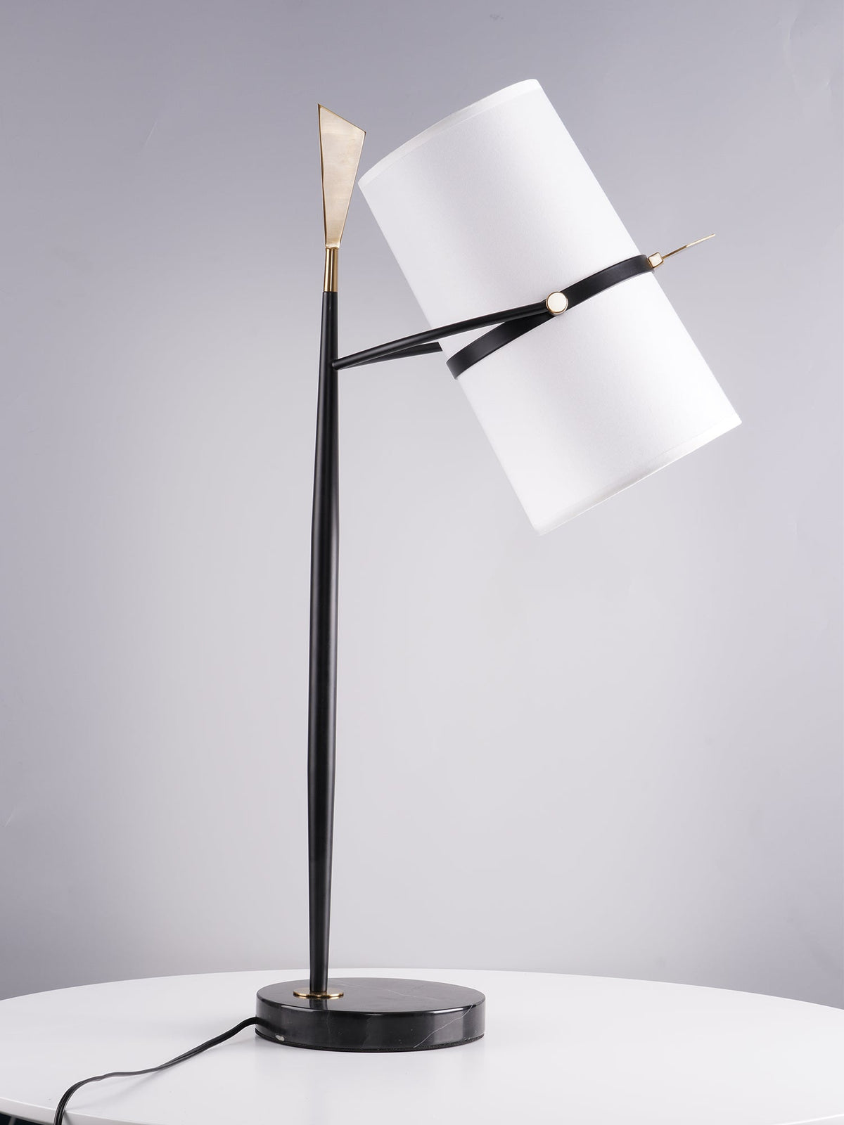 Another angle of this Clérachic black lamp with its shade tilted far over, on a white table, with a gold tip and band against soft grey.