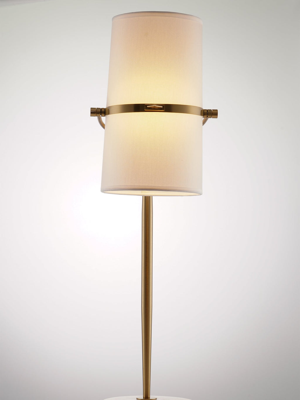 Straight-on view of a Clérachic table lamp lit with warm light, the shade held upright on a gold band and slim gold stem.