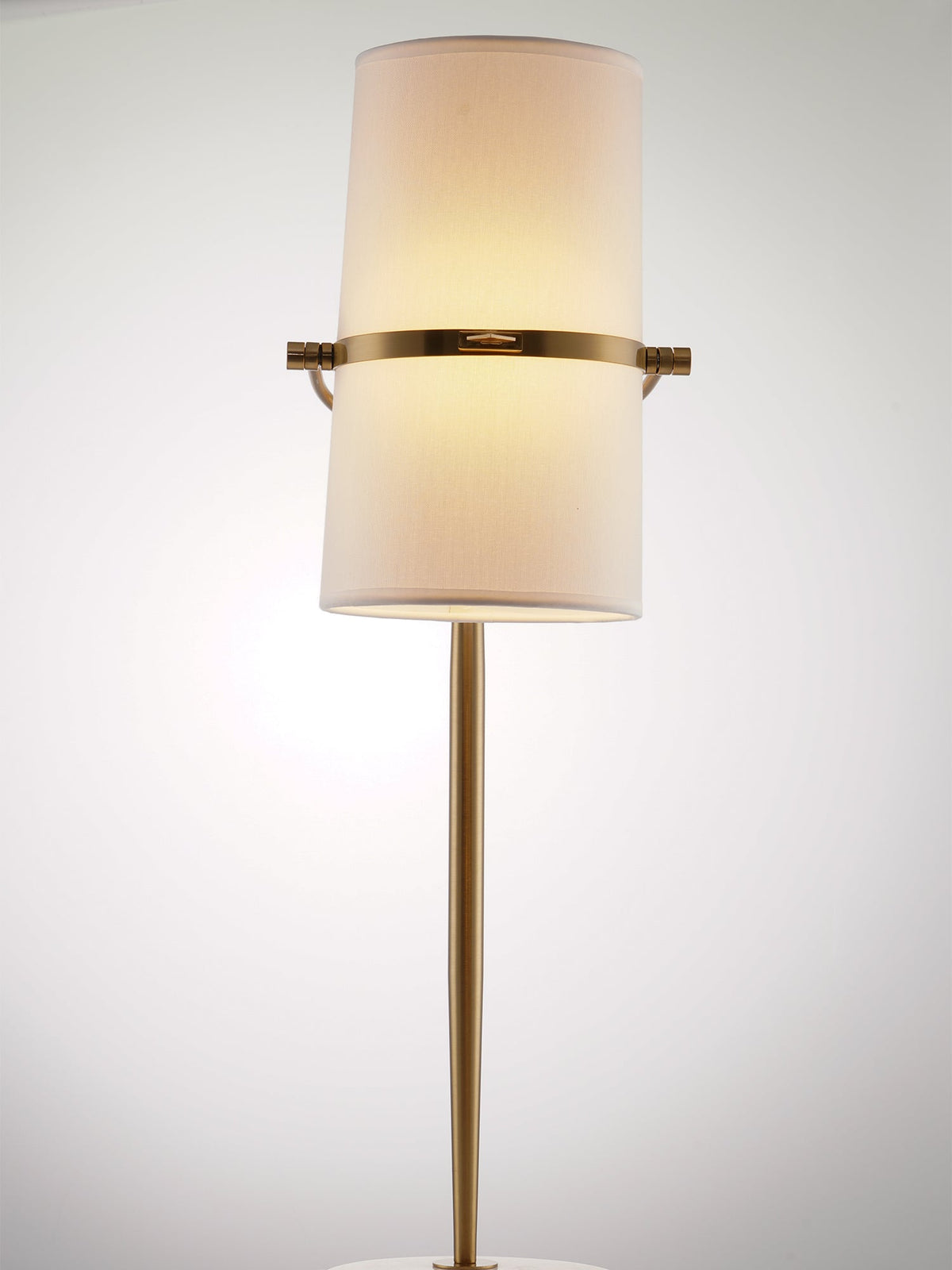 Straight-on view of a Clérachic table lamp lit with warm light, the shade held upright on a gold band and slim gold stem.