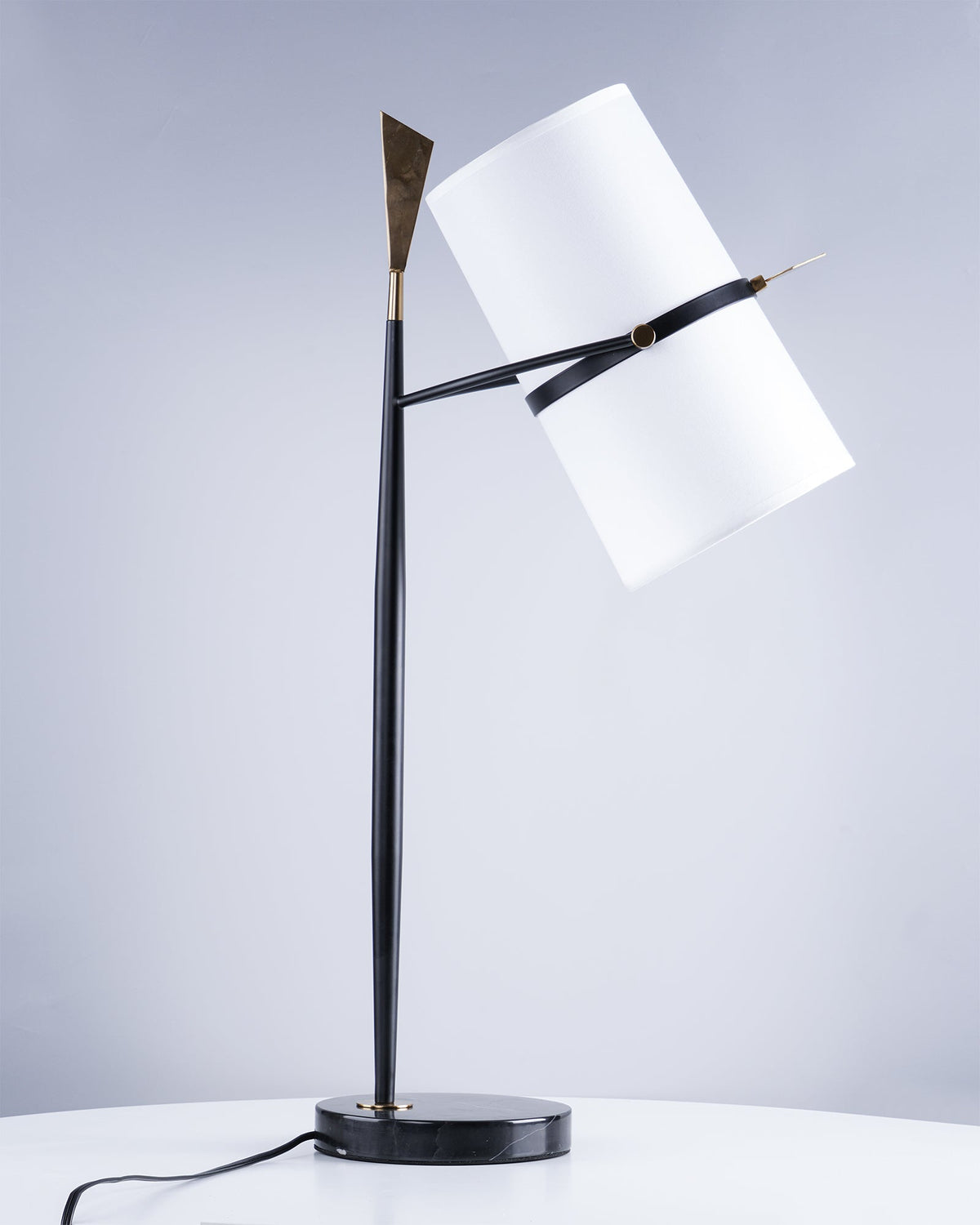 Black Clérachic table lamp on a white table with the shade tilted to the right, against a soft blue-grey backdrop and a trailing cord.