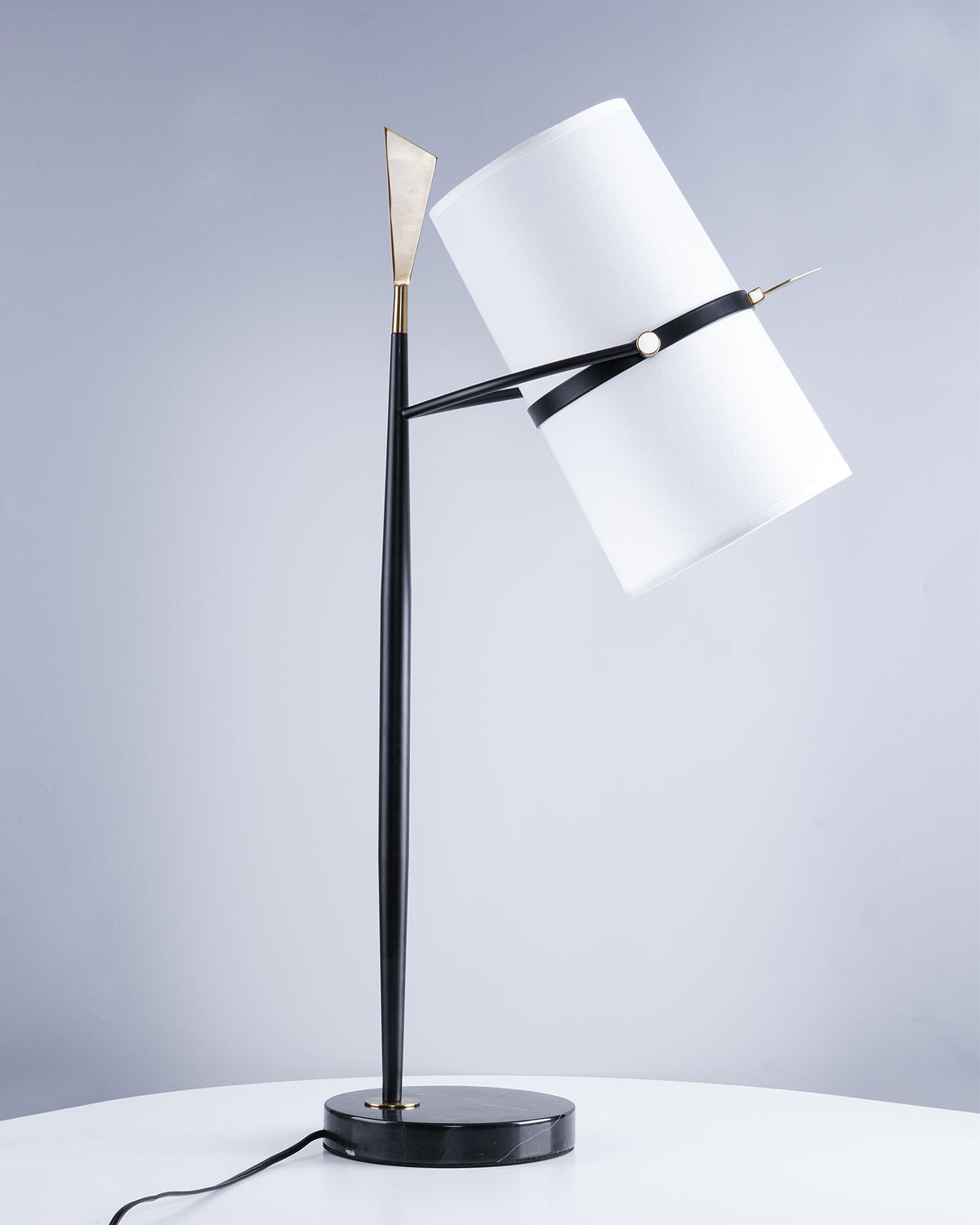 Slightly closer view of this black Clérachic lamp with its white shade tipped down, on a white table with a blue-grey backdrop behind.