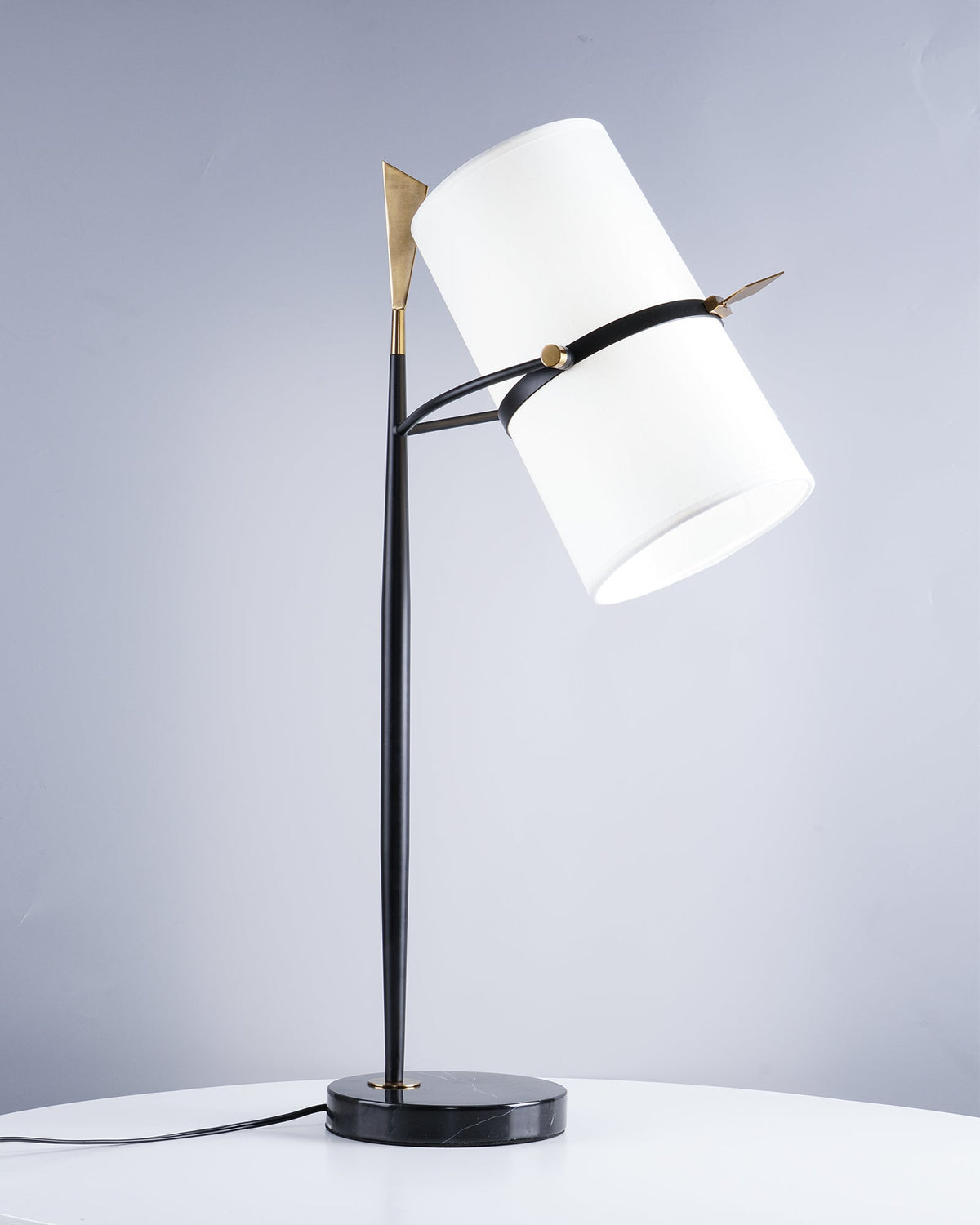 Clérachic table lamp with a black marble base and white shade tilted right, a gold tip at the top, in front of a pale blue-grey wall.