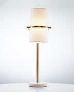 Gold Clérachic table lamp with the shade turned upright and lit warm, on a white marble base against a pale blue-grey backdrop.