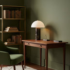 On a walnut writing desk against olive-green walls, this Clérachic table lamp glows beside bookshelves and a green armchair.