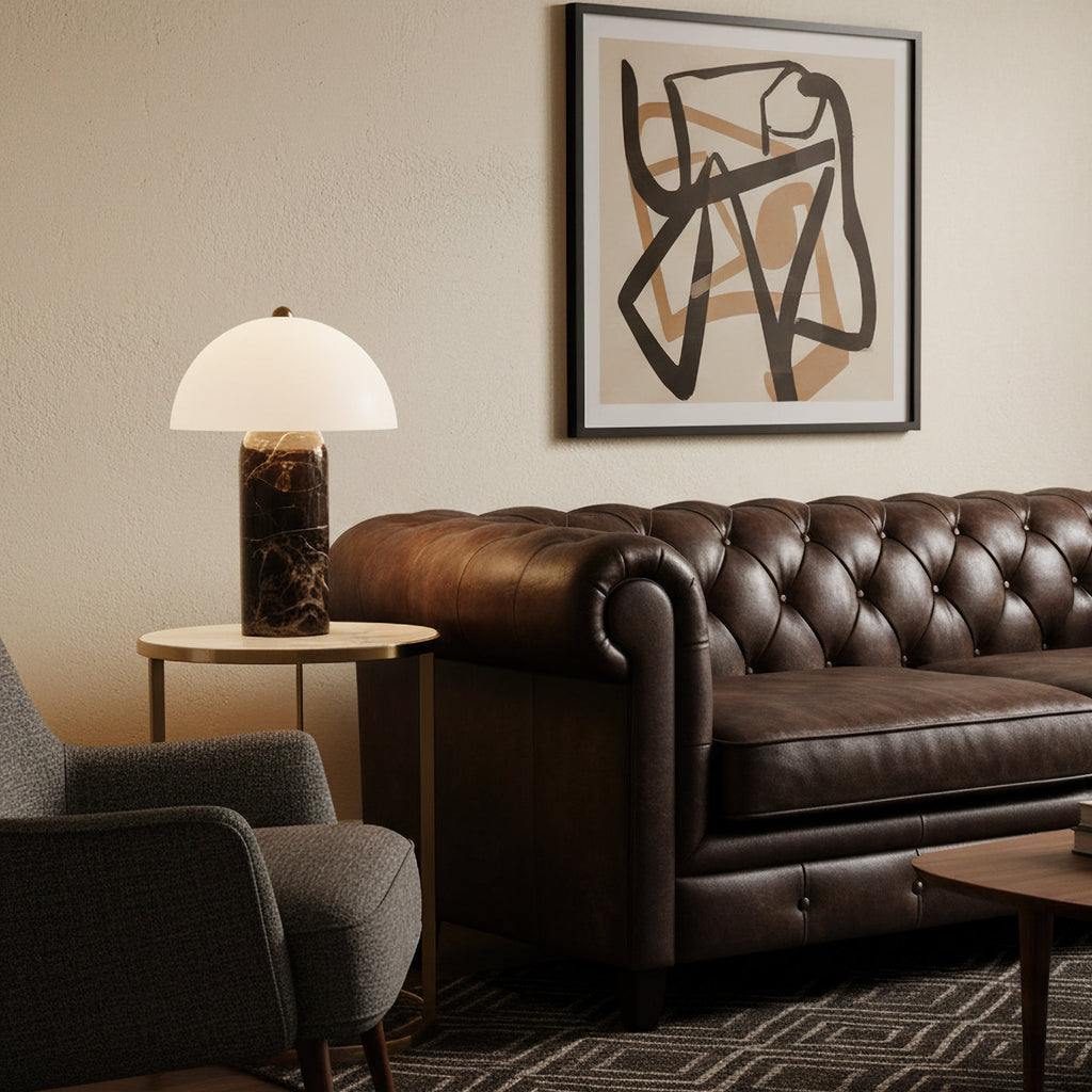Beside a tufted brown leather sofa, this Clérachic table lamp stands on a small brass side table under a framed abstract print.