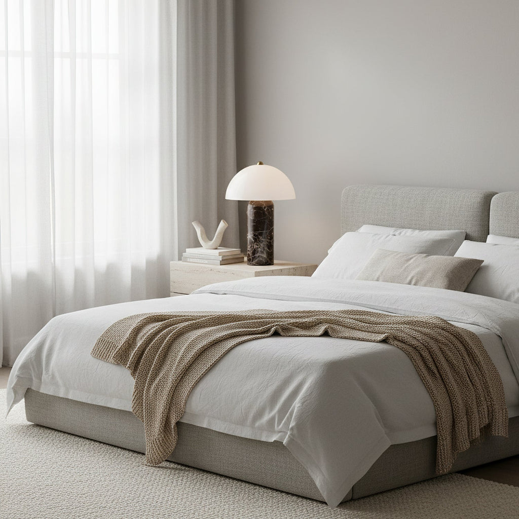 This Clérachic table lamp stands on a pale bedside table beside a white-and-oatmeal bed dressed with a knitted throw.
