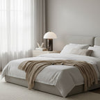 This Clérachic table lamp stands on a pale bedside table beside a white-and-oatmeal bed dressed with a knitted throw.