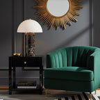 With a sunburst mirror above, this Clérachic table lamp stands on a black side table beside an emerald velvet armchair.