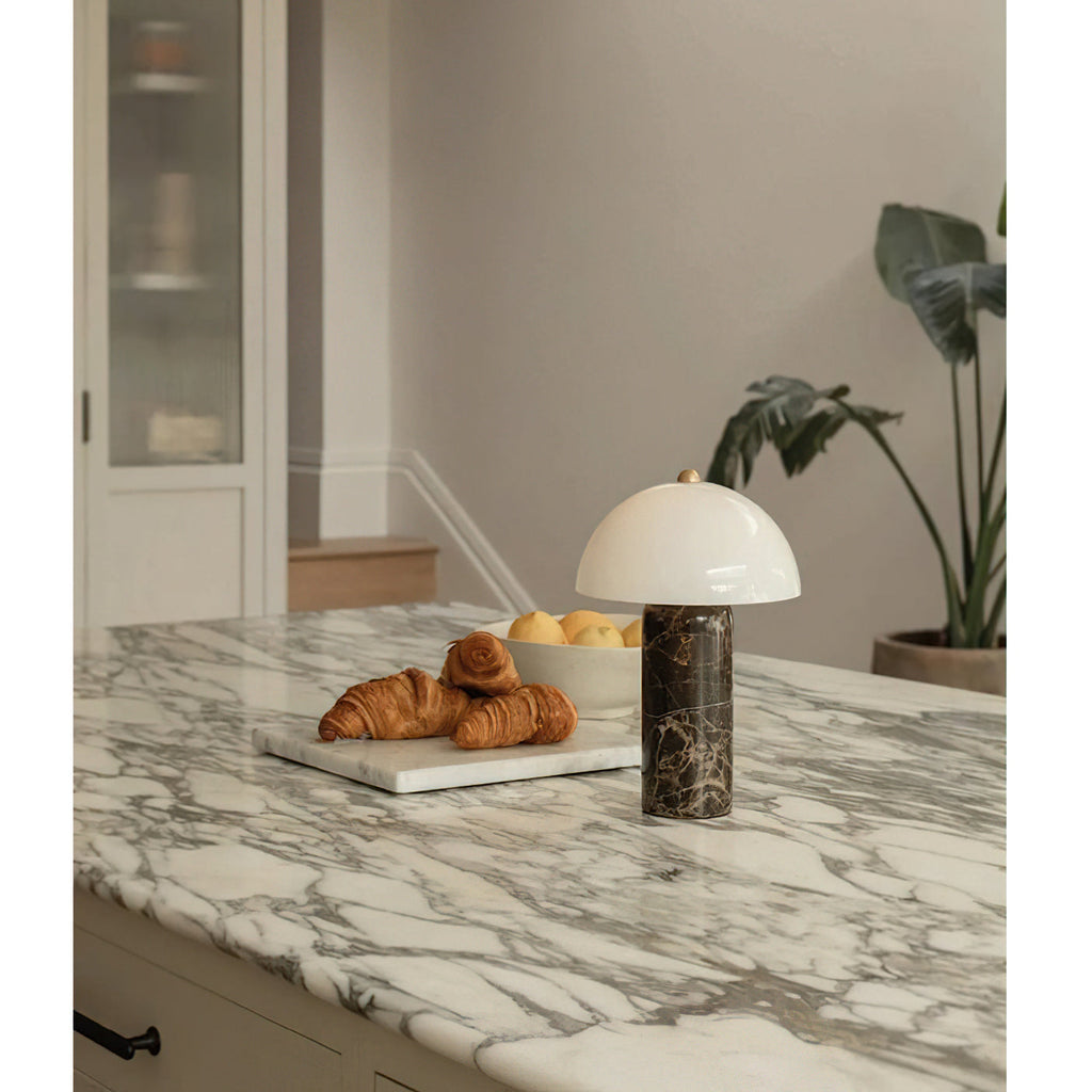 A Clérachic table lamp stands on a marble kitchen counter next to a plate of croissants, with a large plant behind.