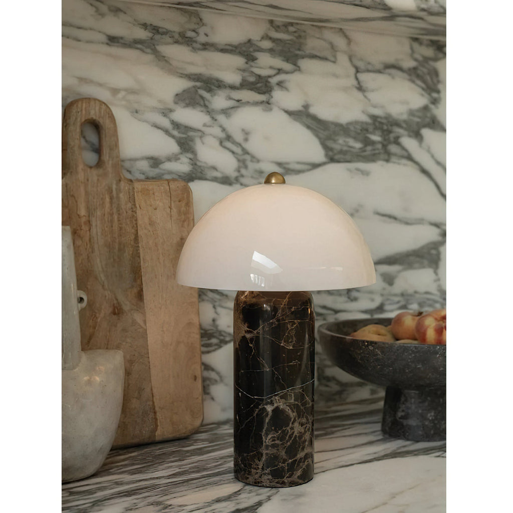 Close view of the domed white shade and veined dark marble base of this Clérachic table lamp, beside a wooden board on a marble splashback.