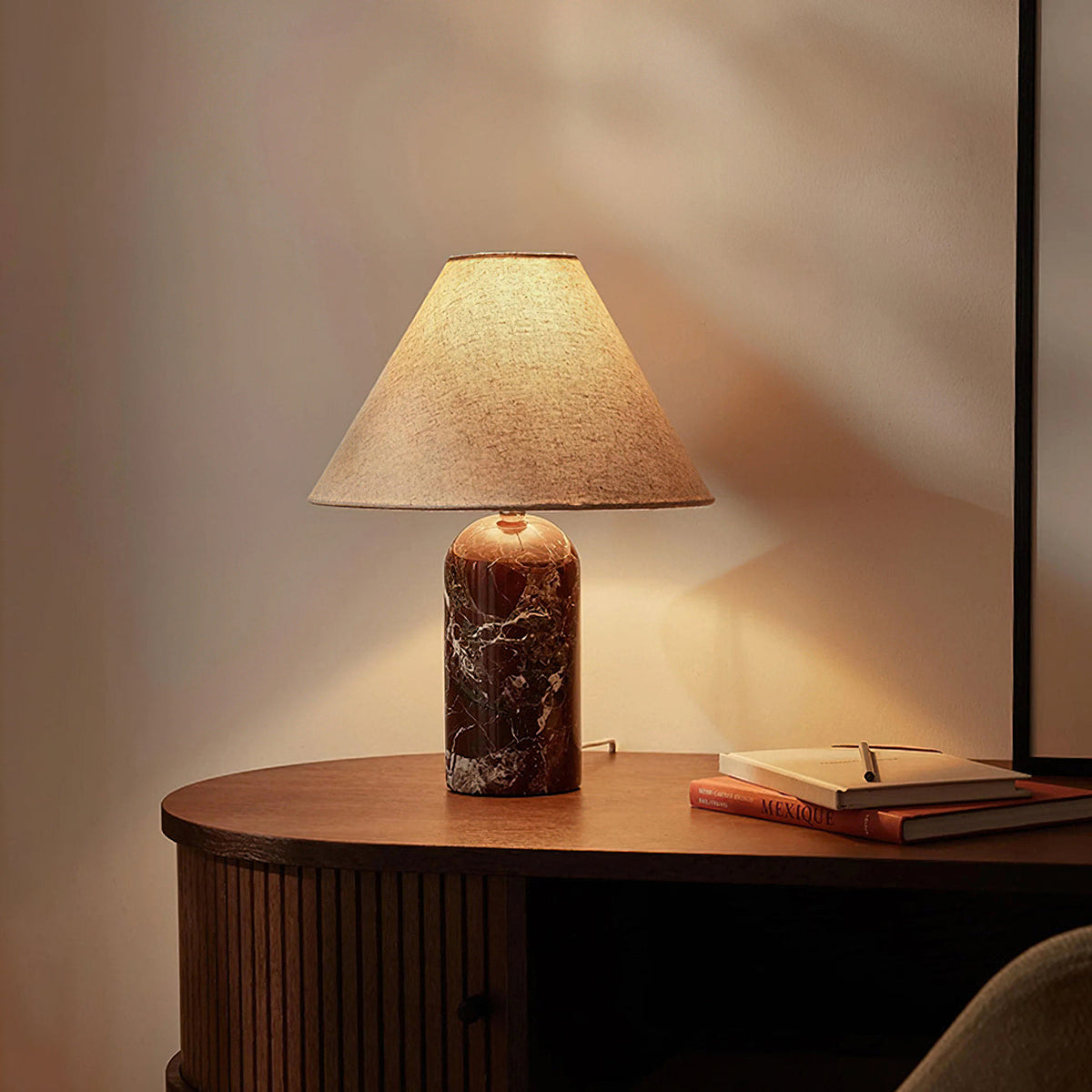 Warm light from a Clérachic table lamp with a dark burgundy marble base, on a curved walnut console with a notebook beside it.