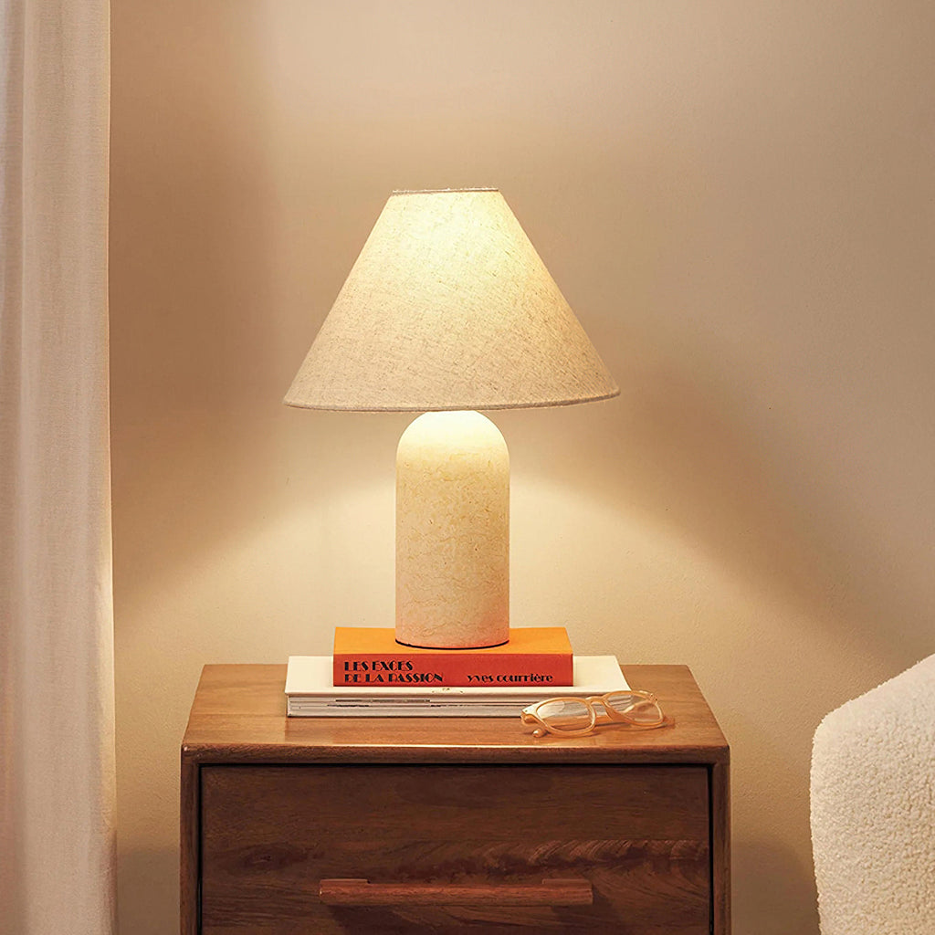 Cream marble base and a glowing linen cone shade on this Clérachic lamp, standing on a stack of books on a bedside chest.