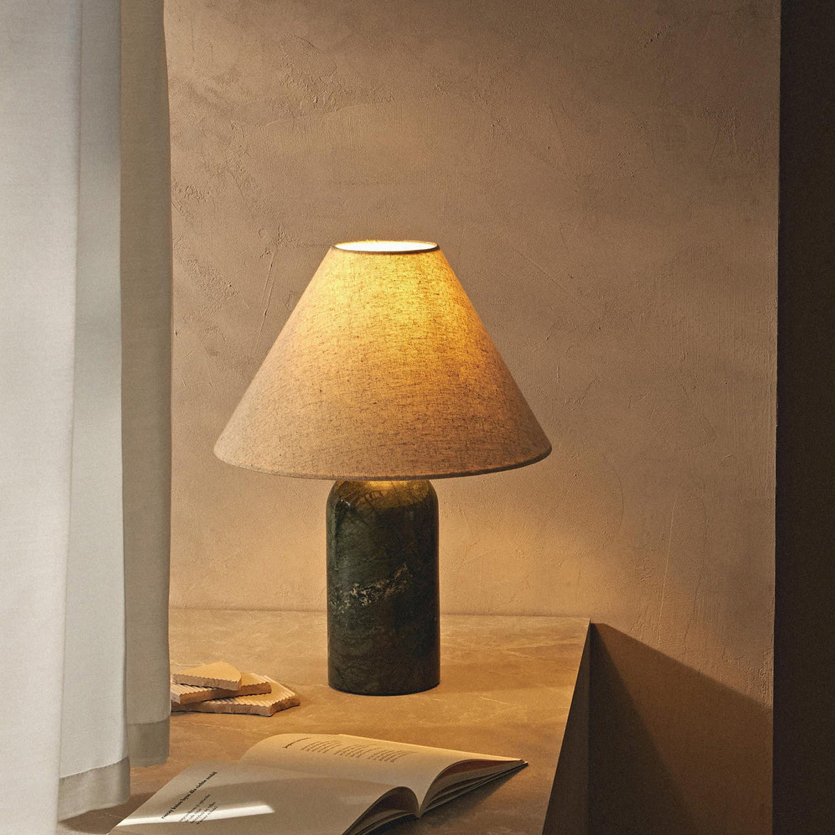 Lit green marble table lamp from Clérachic in a dim corner, its linen shade glowing amber beside a pale curtain and an open book.