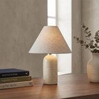 Cream marble base with a pale linen shade, this Clérachic table lamp stands beside a window with a vase of eucalyptus and books.