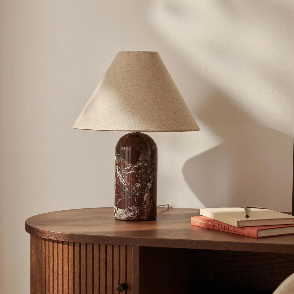 Unlit Clérachic table lamp with a burgundy marble base and natural linen shade, on a ribbed walnut console with two books.