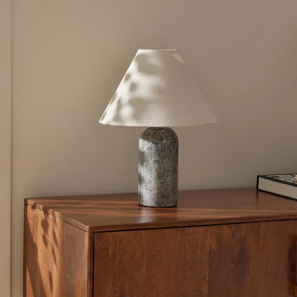 Grey marble base and a white linen shade on this Clérachic table lamp, with dappled sunlight across a walnut chest of drawers.