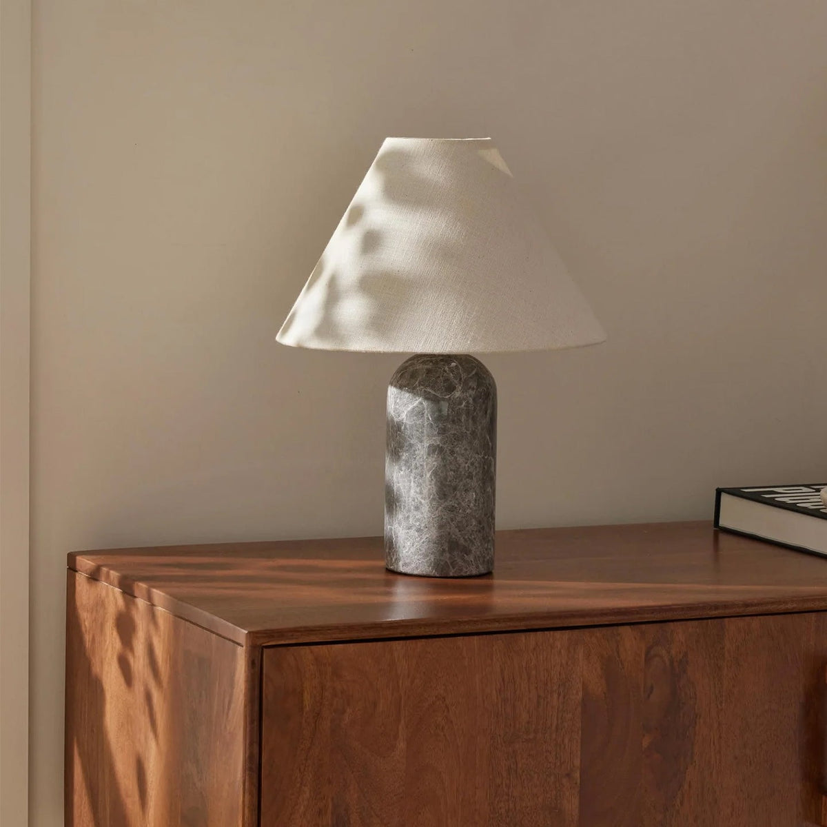 Grey marble base and a white linen shade on this Clérachic table lamp, with dappled sunlight across a walnut chest of drawers.
