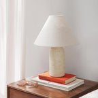 Cream marble table lamp from Clérachic with a pale linen shade, on a bedside table with a stack of books and a pair of glasses.