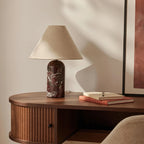 Burgundy marble base on this Clérachic table lamp, with a tan linen shade, on a ribbed walnut console beside a framed print.