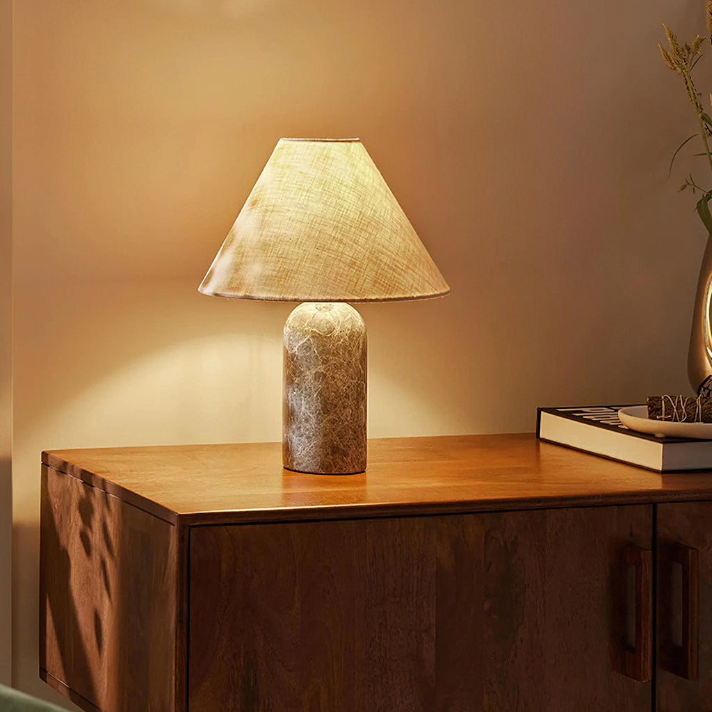 Clérachic table lamp with a lightly patterned grey-brown marble base, lit warmly on a walnut cabinet beside a book and tray.