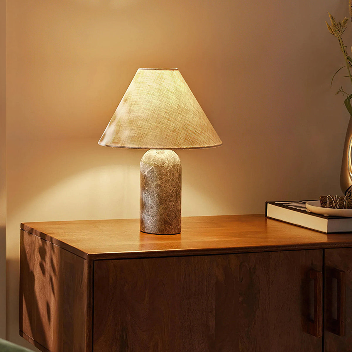 Clérachic table lamp with a lightly patterned grey-brown marble base, lit warmly on a walnut cabinet beside a book and tray.