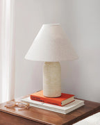 Beige marble table lamp from Clérachic, standing on two books on a walnut side table in soft daylight beside a sheer curtain.
