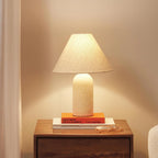 A Clérachic table lamp with a cream marble base glows on a bedside table, with a stack of books and glasses below the shade.