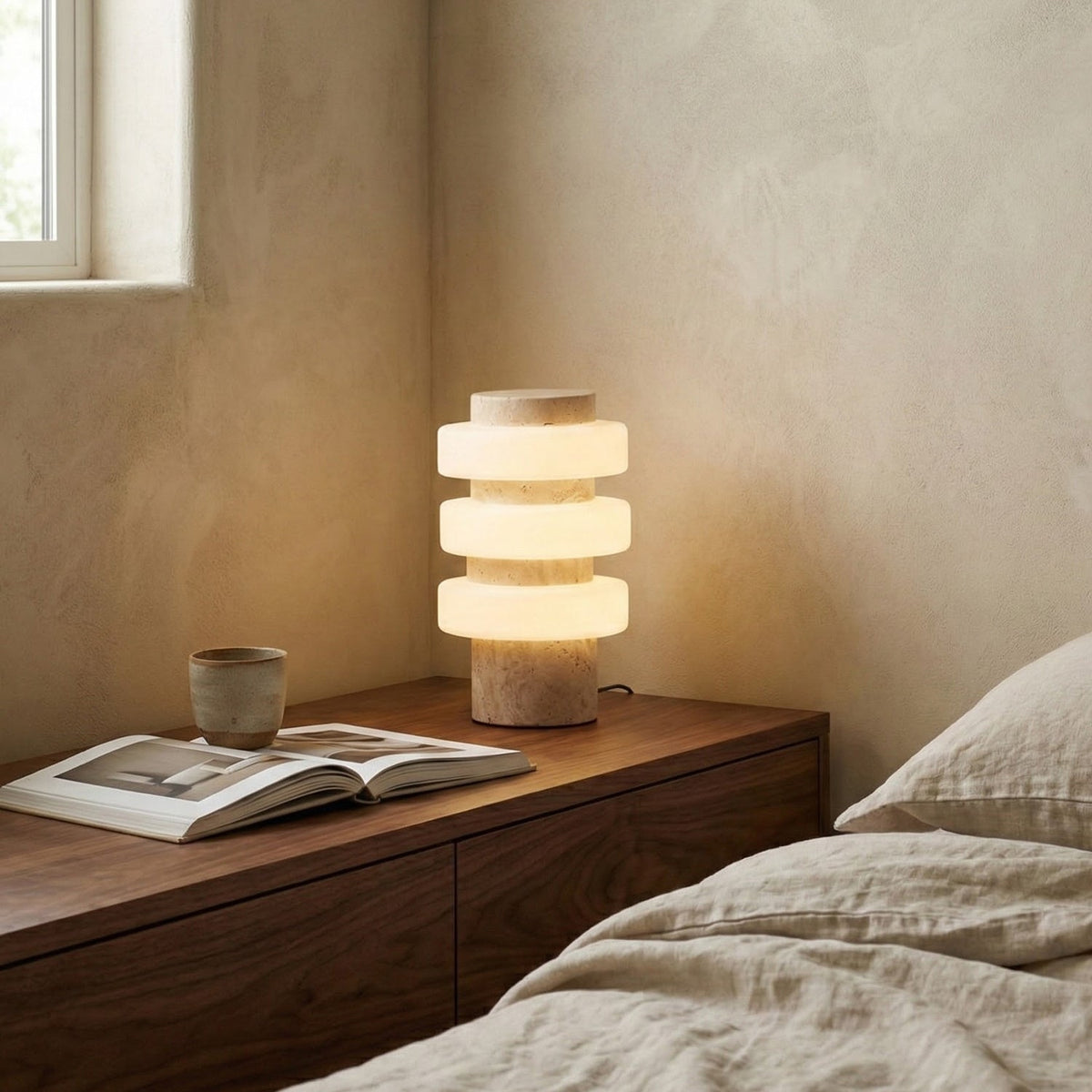A lit Clérachic table lamp with three glowing glass discs on a beige stone base, on a walnut bedside unit beside an open book and a ceramic cup.