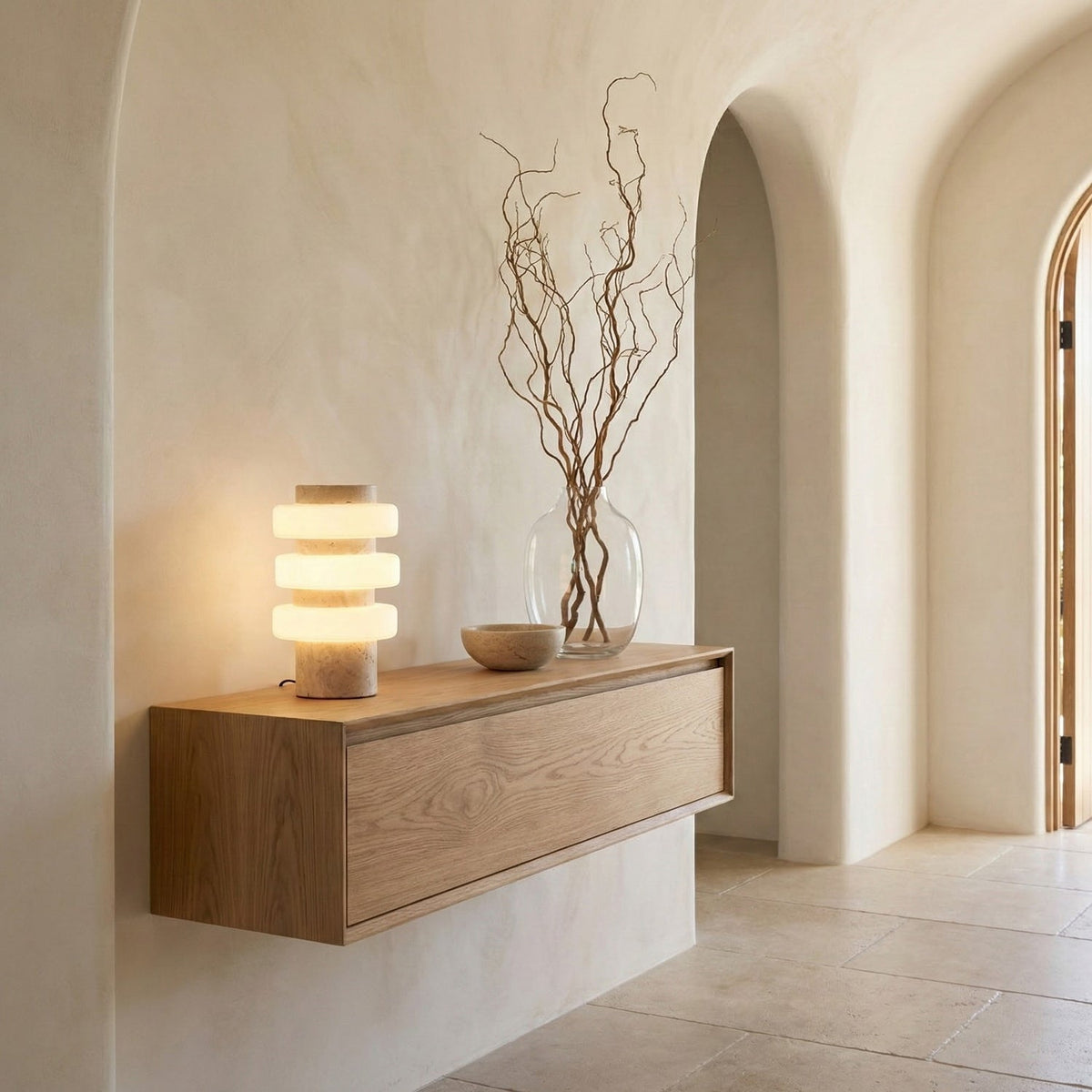 A small Clérachic table lamp glows on a floating oak shelf in an arched, plastered hallway, with bare branches in a glass vase beside it.
