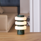 A Clérachic table lamp in the green and white finish glows on a wooden table, with a cream sofa softly blurred behind it.