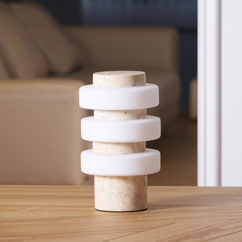 A slightly different angle of the unlit Clérachic table lamp, its white glass discs and beige stone column on a herringbone wooden table.